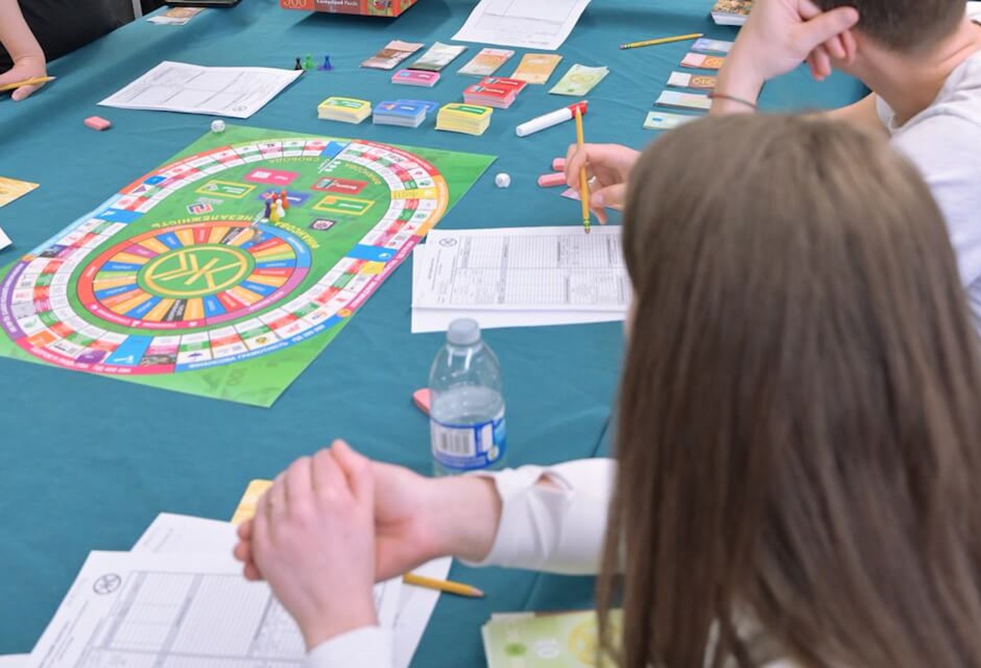 A Life Capital tournament session in progress: the strategy board, asset cards, play currency, and player ledgers on the emerald baize.